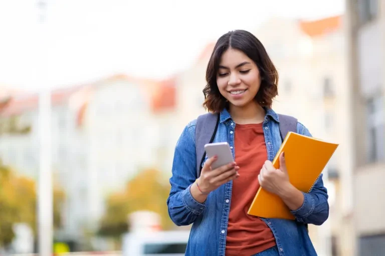 Smiling young Middle Eastern student walking with a smartphone and binders on a city street – Cheerful young Arab woman texting on her mobile phone while heading home after class.