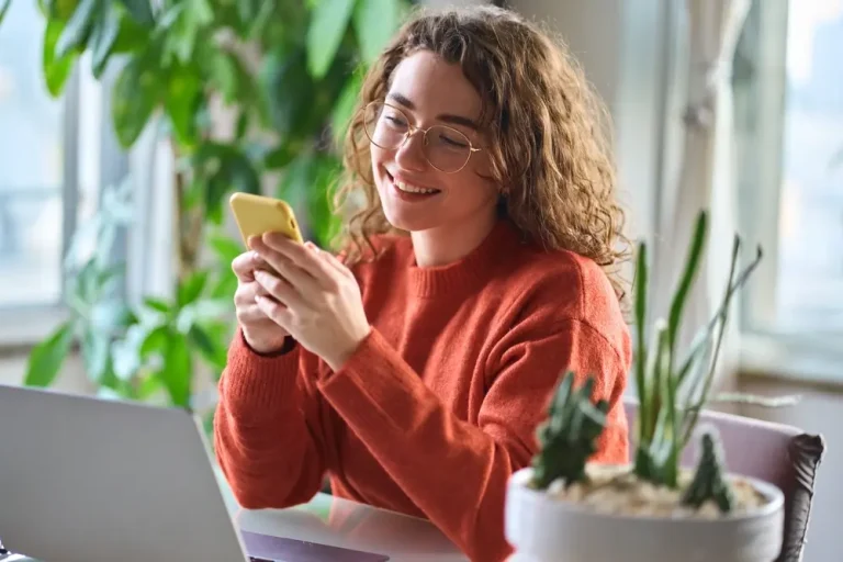 Happy, smiling young woman sitting at a table holding a smartphone – using modern mobile phone technology, looking at her phone while working.
