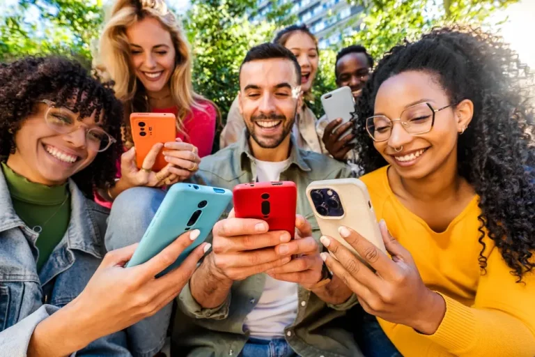 Group of young people using a smartphone outdoors – Happy friends laughing together while watching a funny video on a social media platform – Modern technology and lifestyle concept.